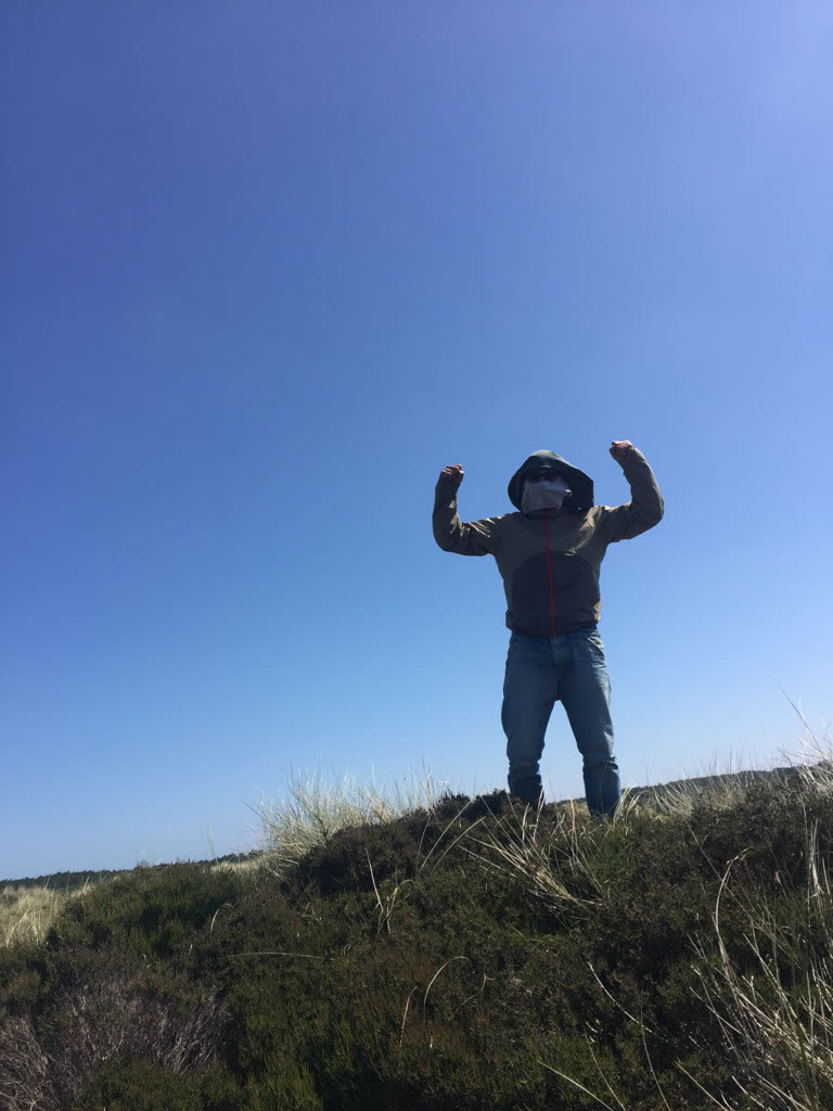 Iemand staat met opgeheven armen op een duin onder een blauwe lucht